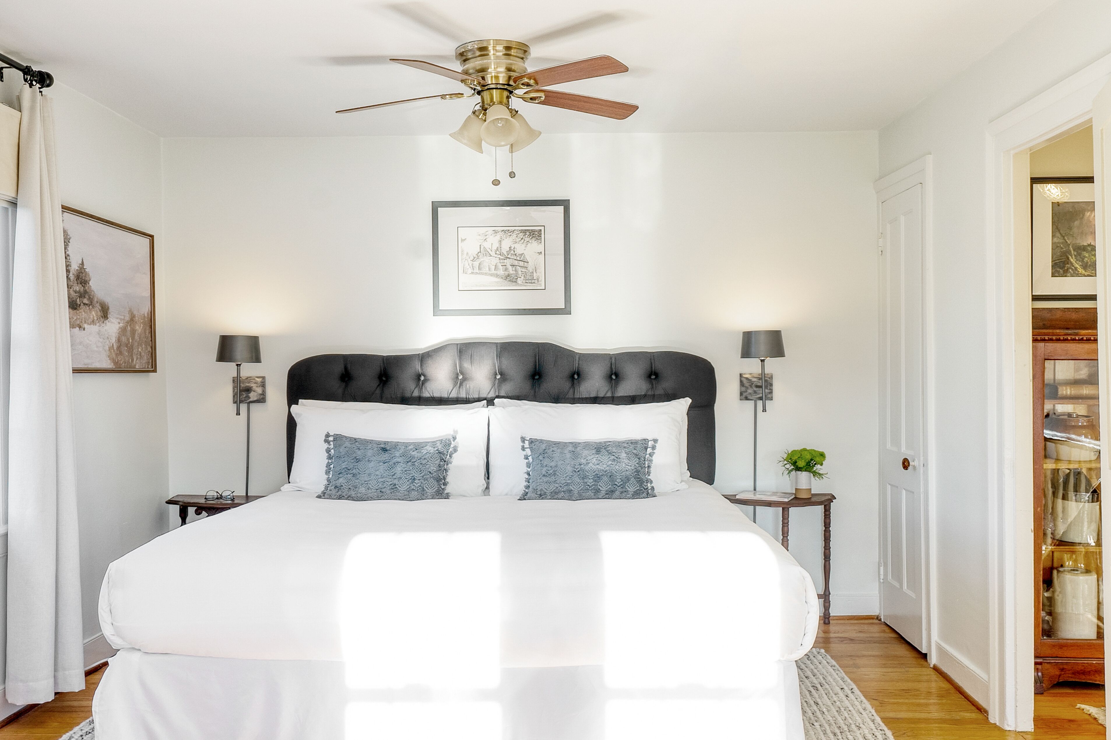 A bedroom with a large bed, a ceiling fan, a framed picture on the wall, and a nightstand with a lamp.
