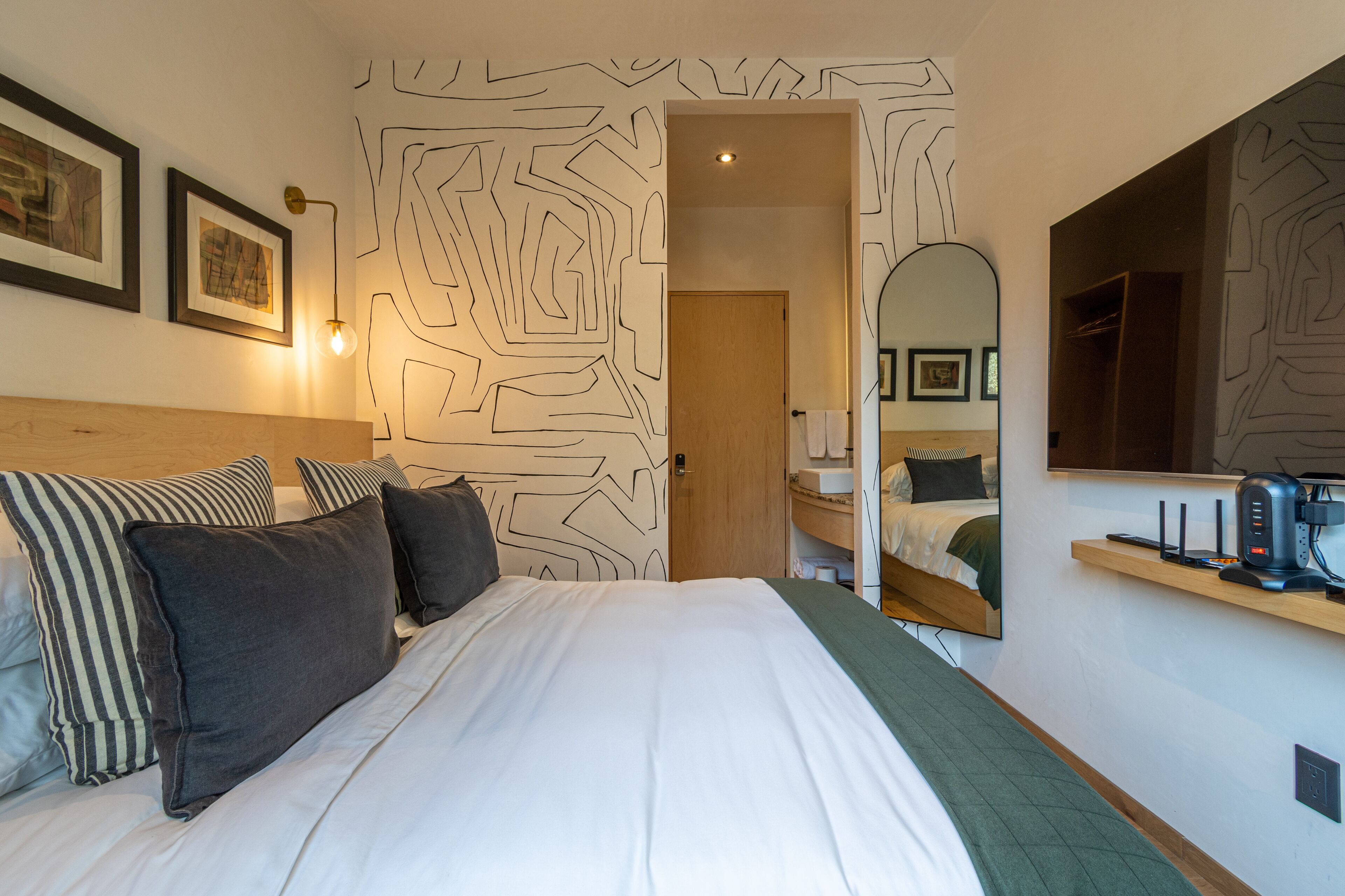 Una habitación de hotel moderna con una cama grande, un cabecero de madera, un espejo, una estantería con cafetera y arte abstracto en la pared.