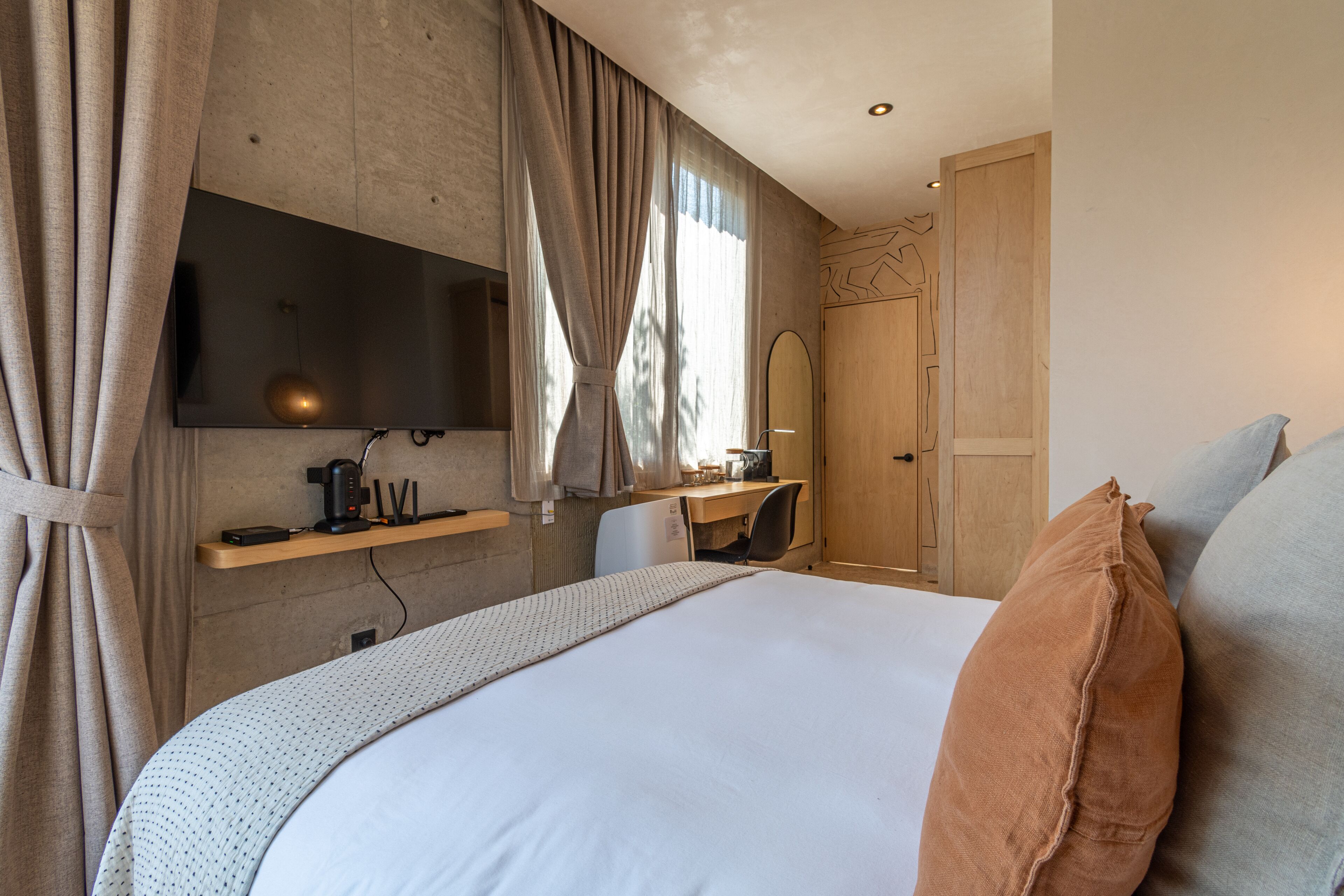 A modern hotel room with a large bed, a desk with a chair, a flat-screen TV mounted on the wall, and a wooden door.