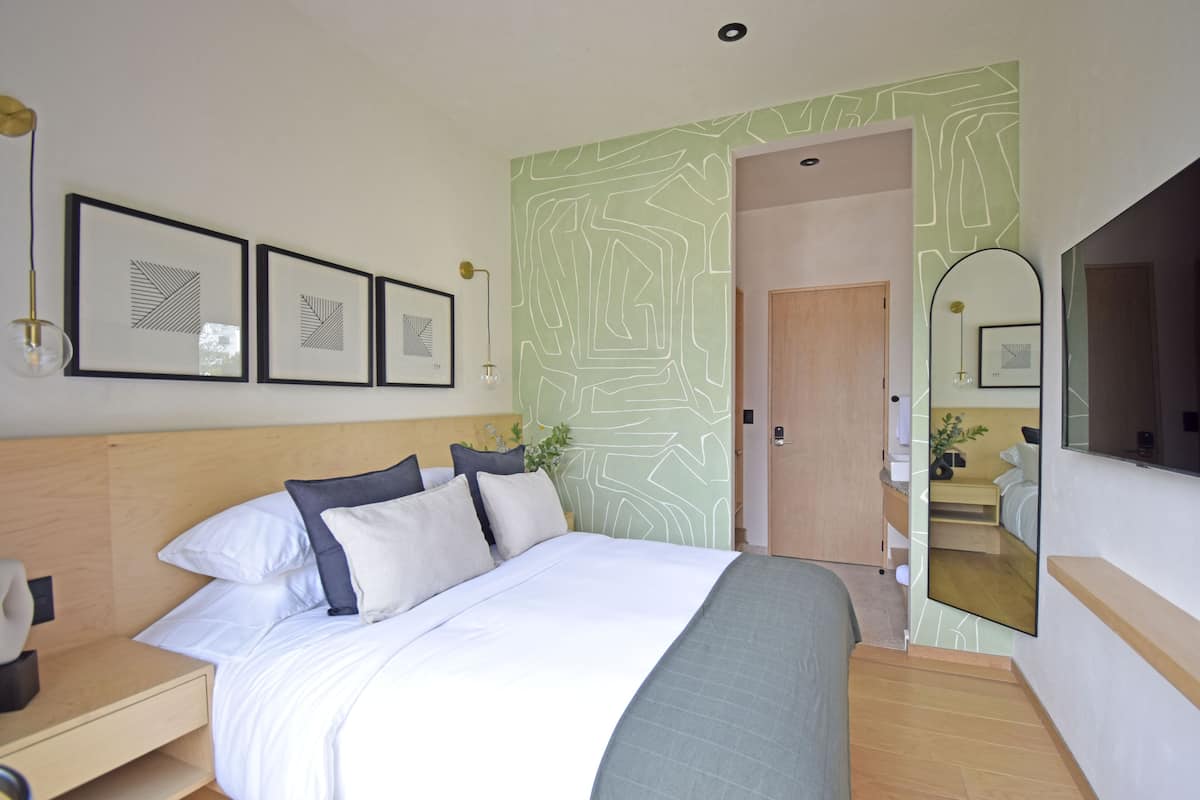 A modern hotel room with a large bed, a bedside table, a mirror, and framed artwork on the wall.