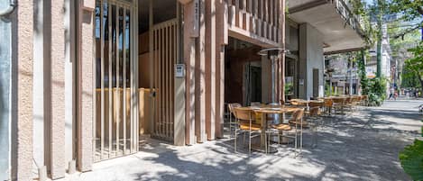 Un café al aire libre con mesas y sillas, la entrada de un edificio con un número, y una vista de la calle.