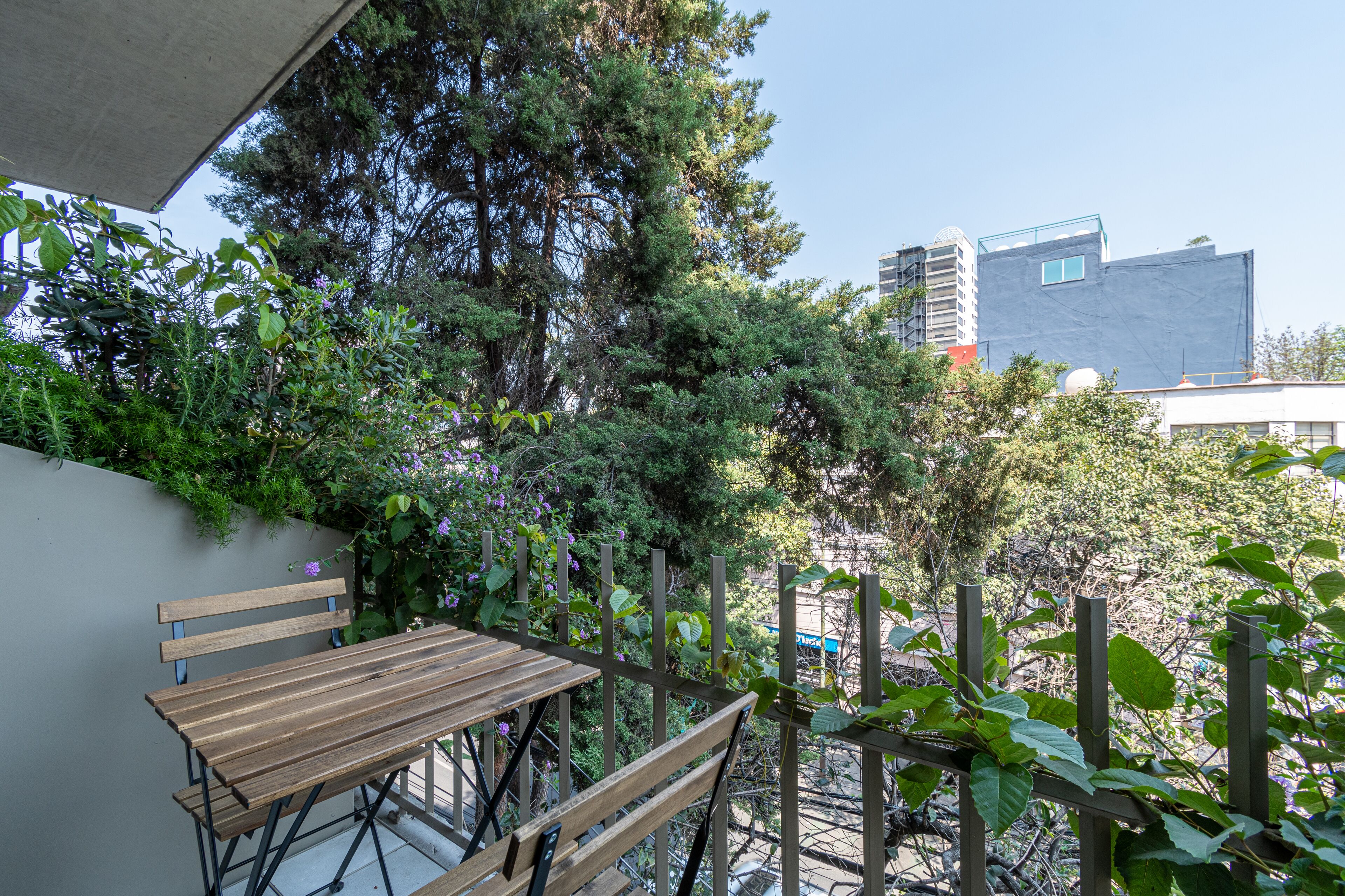 Un balcon avec une table et des chaises en bois, entouré de verdure et offrant une vue sur les bâtiments en arrière-plan.