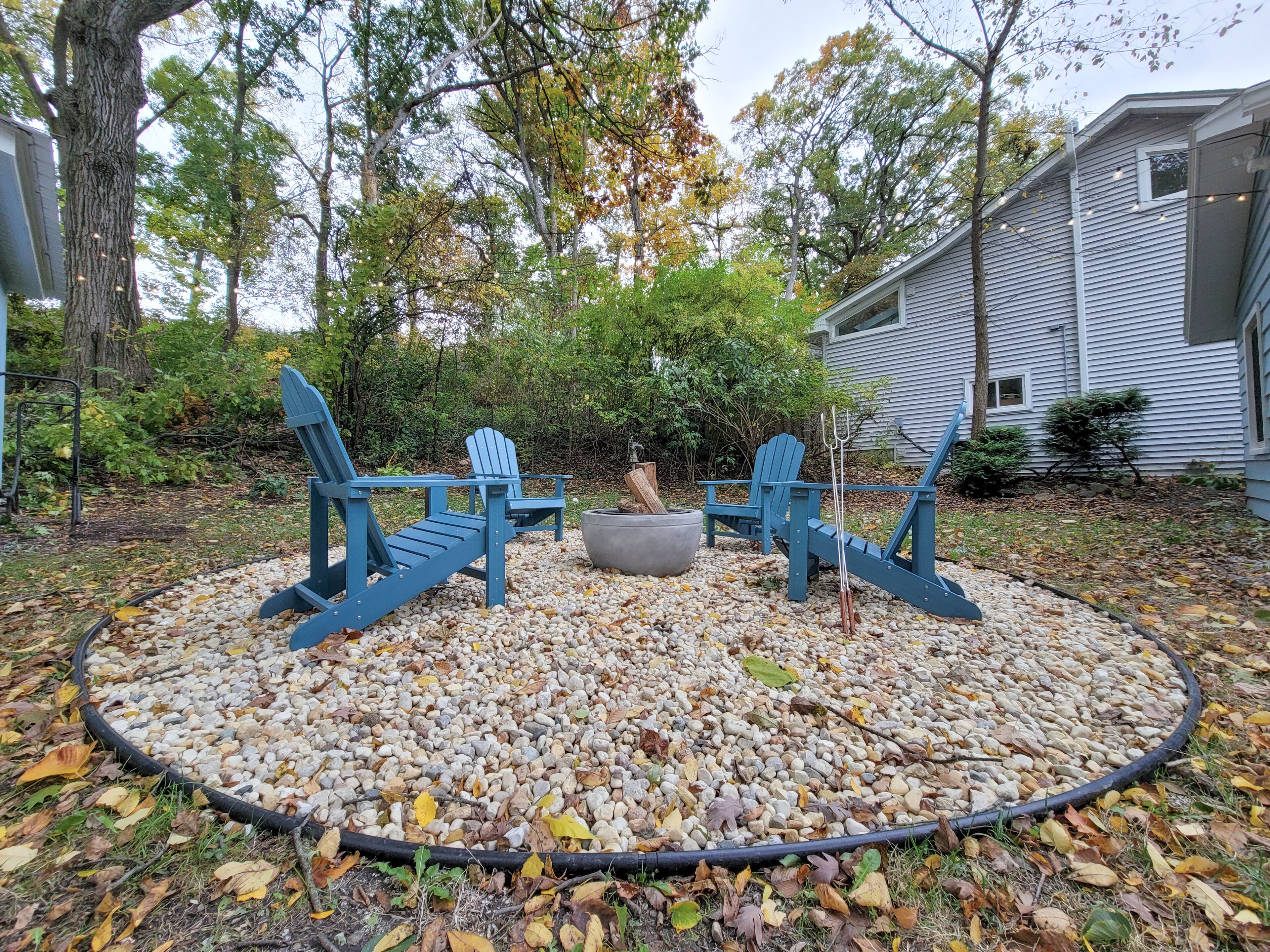 A circular seating area with Adirondack chairs and a fire pit, surrounded by pebbles and trees.
