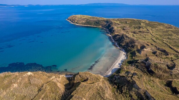 An aerial view of a coastal landscape with a beach, grassy hills, and a clear blue sea.