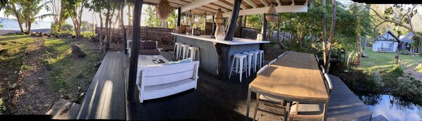 A covered outdoor seating area with a bar, wooden deck, and a view of a house and trees.