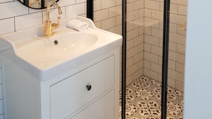 Un baño moderno con un lavabo blanco, detalles dorados, un espejo grande y una zona de ducha con mampara de vidrio.