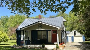 A small, single-story house with a dark blue exterior, a red door, and a chimney. It has a porch with outdoor seating and a small garden area with flowers. - Lexington Stays. Close to town and beach!  (Lexington)