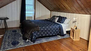 A bedroom with a sloped ceiling, a bed with navy bedding, a small bedside table, and a window with blinds. - Lexington Stays. Close to town and beach!  (Lexington)