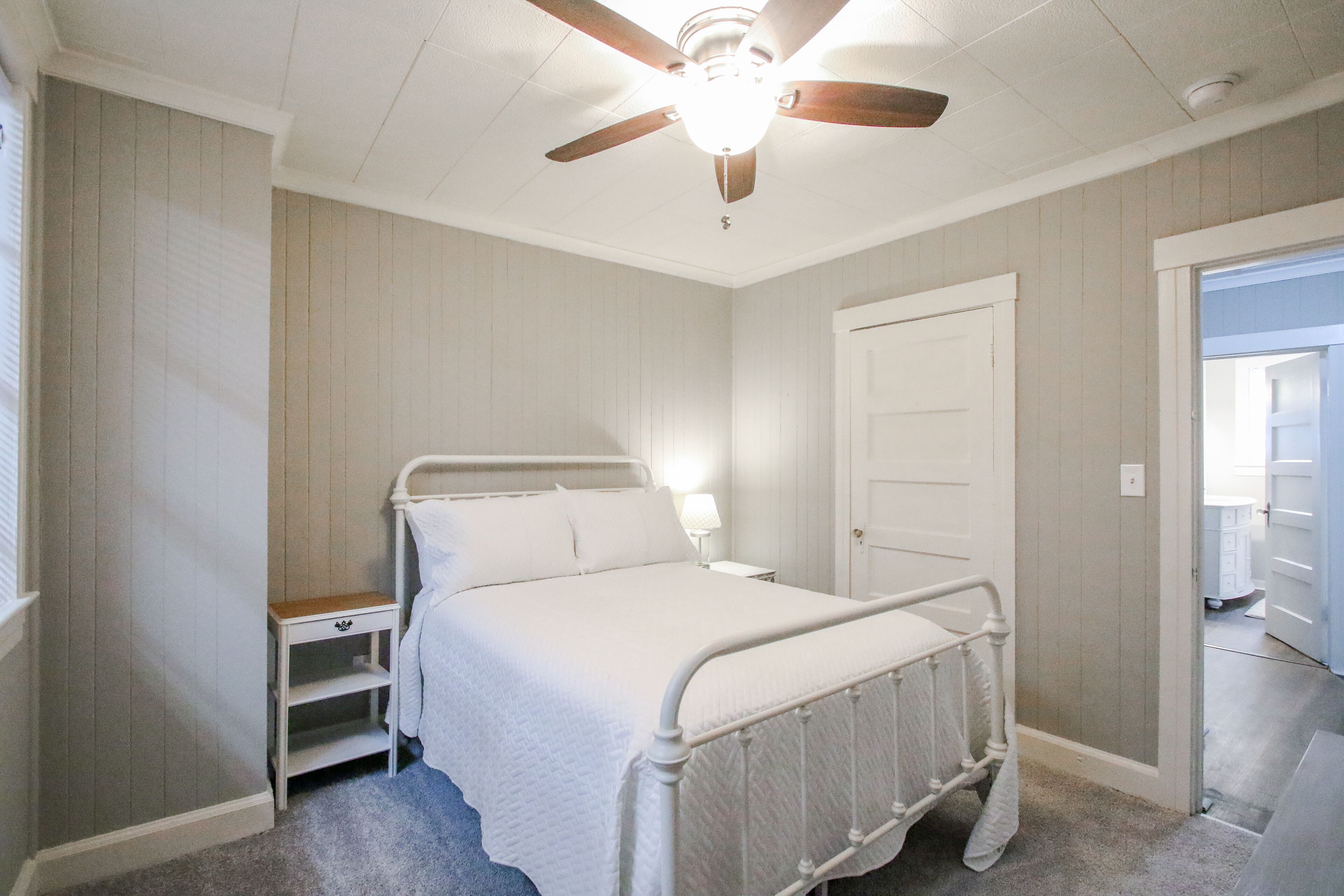 A bedroom with a single bed, a nightstand, a ceiling fan, and a door leading to another room.