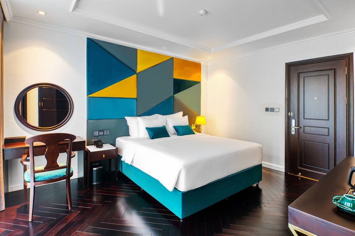 A hotel room with a bed, a desk, a chair, a bedside table, and a door.
