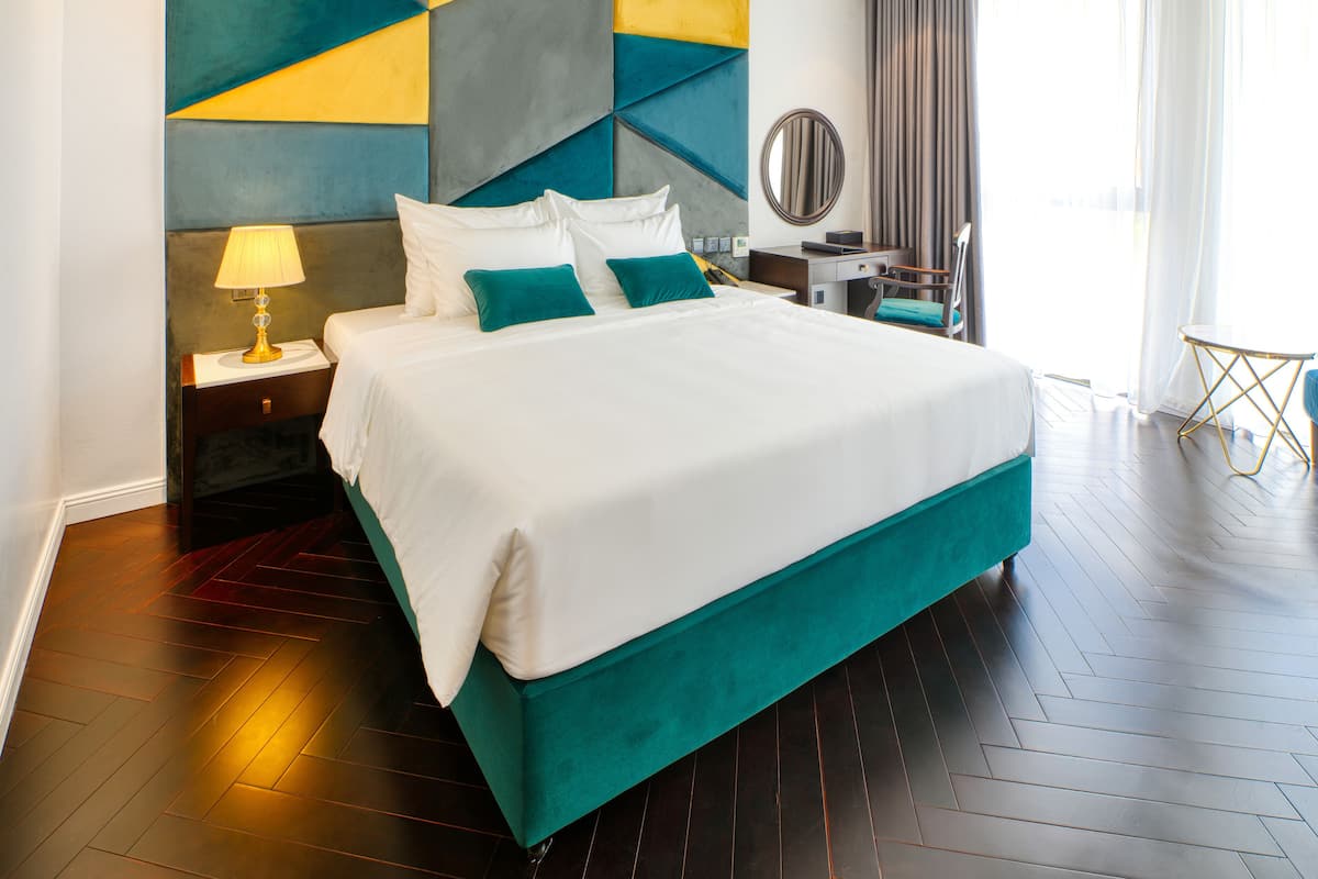 A hotel room with a large bed, a nightstand with a lamp, a desk with a chair, and a view of the outdoors.