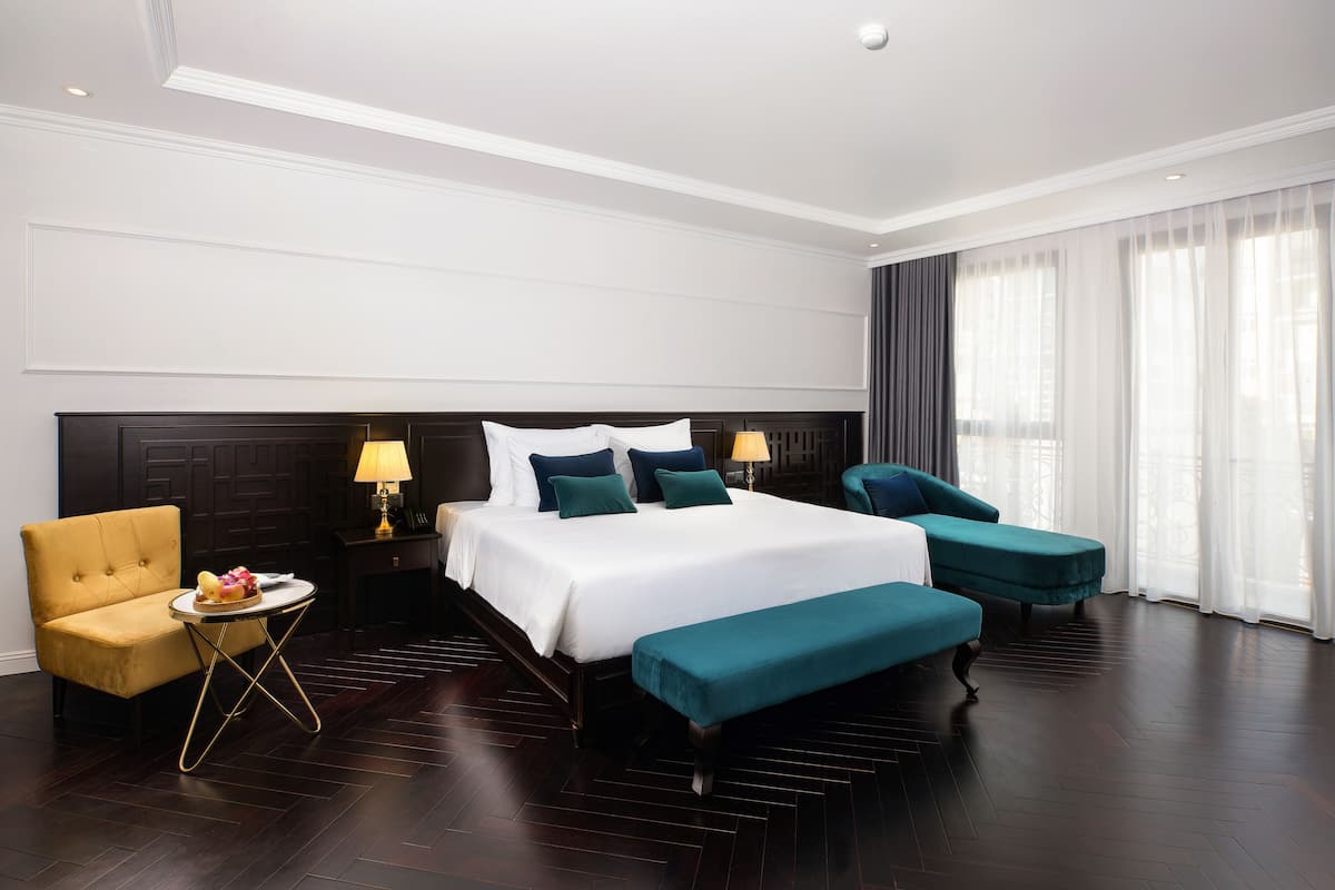 A hotel room with a large bed, a sofa, a small table with a lamp, and a bench.