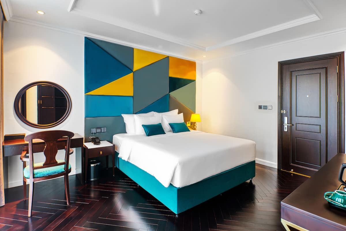 A hotel room with a large bed, a desk with a chair, a bedside table, and a wall with geometric patterns.