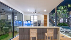 A modern kitchen with a central island, two bar stools, and a view of a pool area. - Villa Ananta - 3 Bedroom (Hikkaduwa)