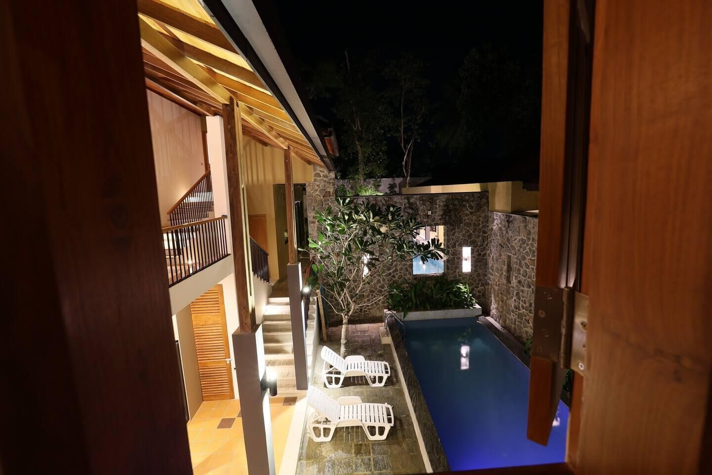 A nighttime view of a modern outdoor swimming pool area with a stone wall, a wooden deck, and a staircase leading to an upper level.