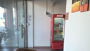 Una zona de estar con sillas de madera, un acceso por puerta de vidrio, un ventilador fijado a la pared y un refrigerador rojo Coca-Cola.