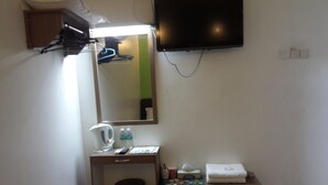 Una habitación pequeña y funcional con escritorio, silla, espejo, televisor montado en la pared y un mueble auxiliar.