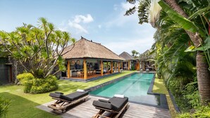 A poolside area with lounge chairs, a thatched-roof structure, and lush tropical vegetation. - Villa Wolfe by Villa Finder (Seminyak)