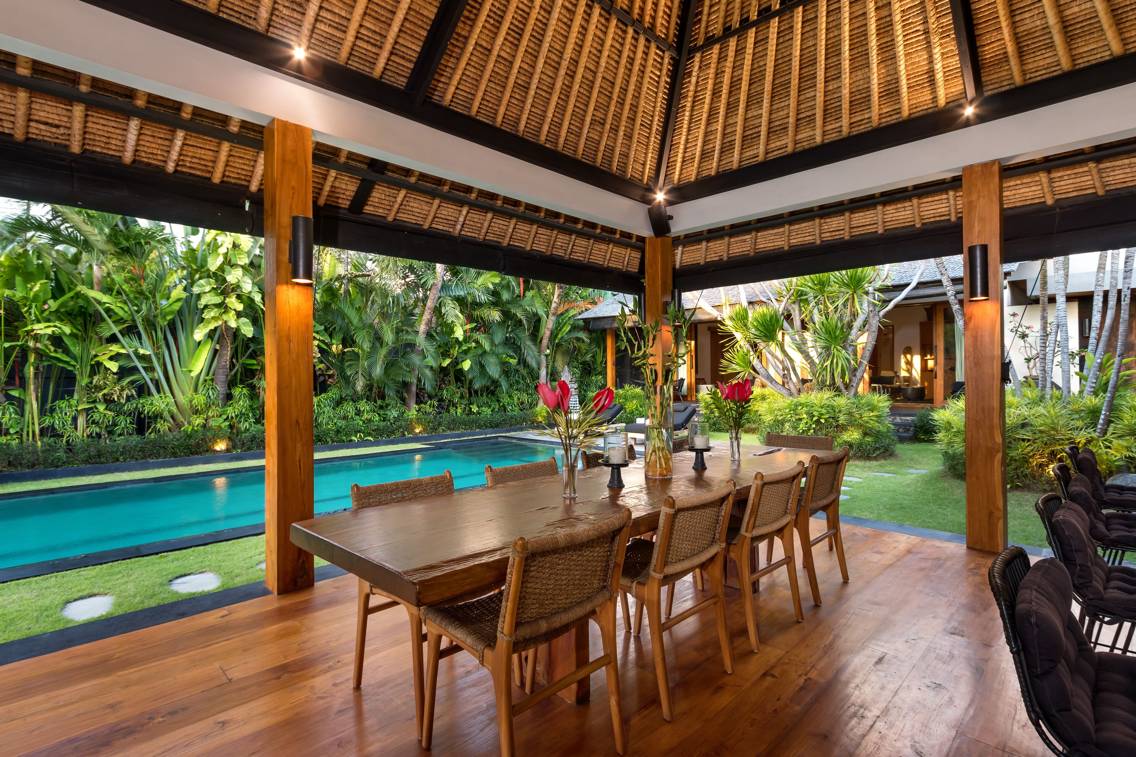 An outdoor dining area with a pool, surrounded by lush greenery and a thatched roof structure.