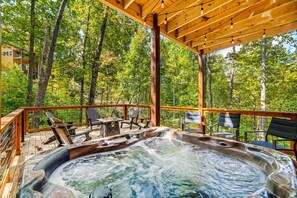 Relax on the elevated deck with a hot tub at its center, surrounded by seating and tables, offering a view of the surrounding forest.