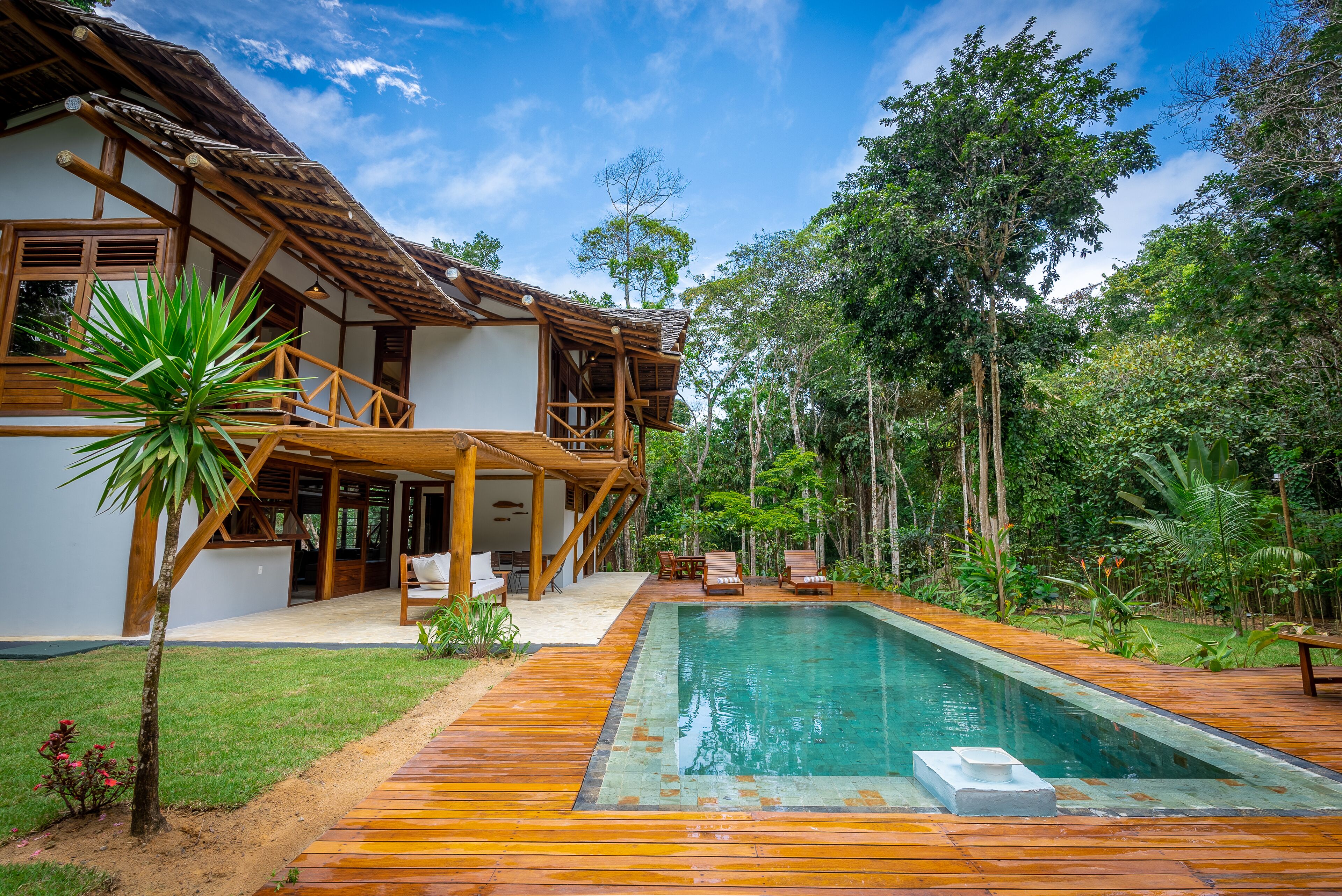 Una terraza de madera que conduce a una piscina, rodeada de exuberante vegetación y una casa con techo de paja.