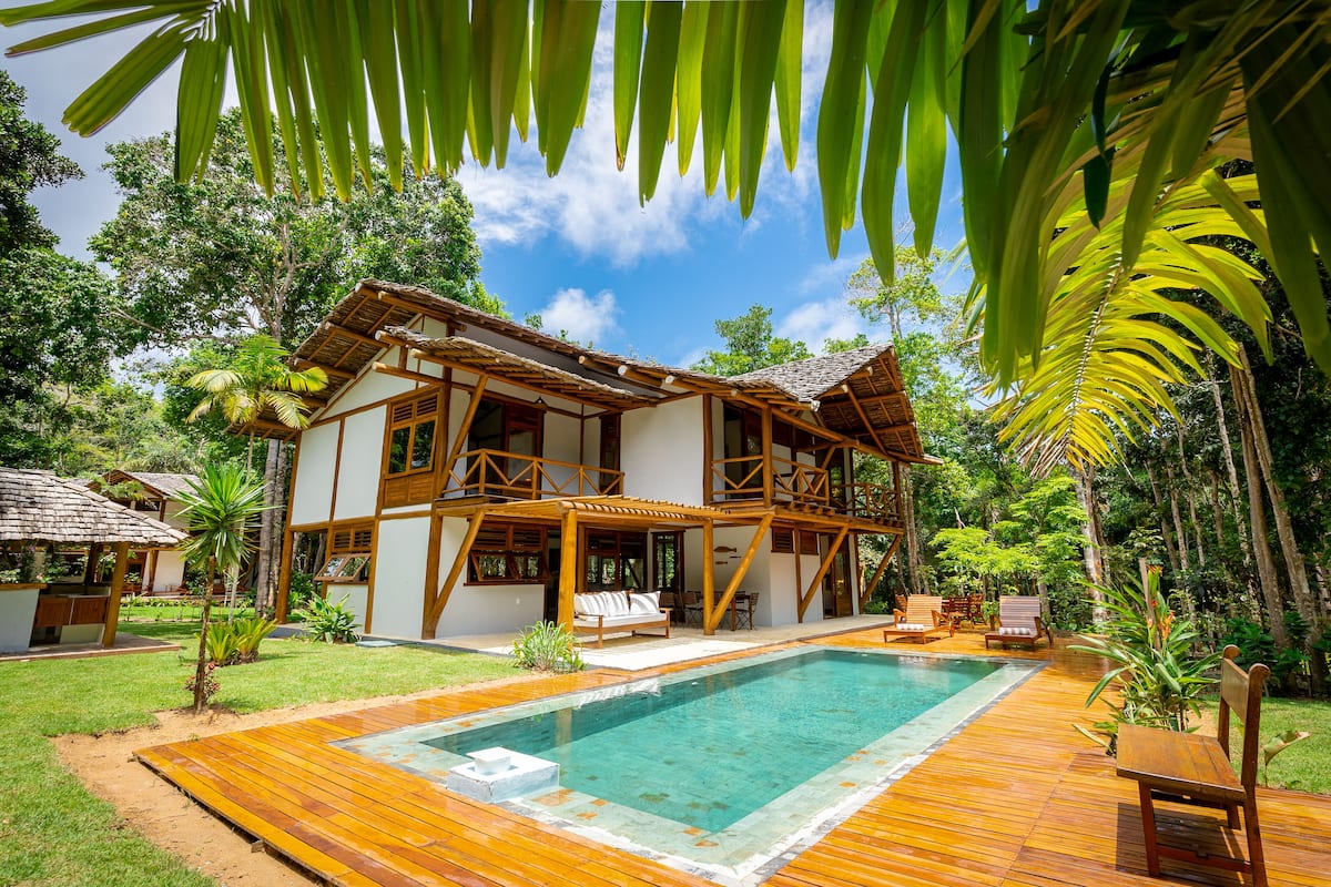 Una casa de madera de dos pisos con piscina, rodeada de vegetación exuberante y palmeras.