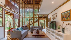 Un salón amplio con techo alto, vigas de madera y ventanas grandes. Hay dos sofás, una mesa de centro y un televisor sobre un mueble blanco.
