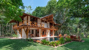 Una casa de madera de dos pisos con un balcón, rodeada de exuberante vegetación y una piscina.