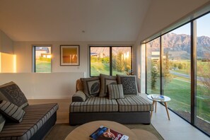 A modern living room with a large window offering a view of a golf course and mountains. There is a sofa with patterned cushions and a small side table with a magazine.