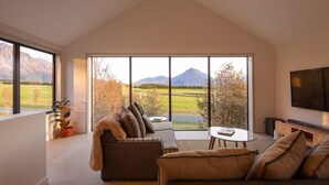 A spacious living room with a high ceiling, large windows offering a view of mountains, a cozy seating area with a sofa and armchairs, and a flat-screen TV mounted on the wall. - Beautiful 3 bedroom home | Hot Tub | Stunning View (Jacks Point)