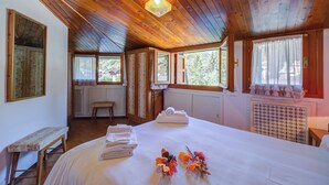 Una stanza con soffitto in legno, un letto con copripiumino bianco, una panca, una finestra vista alberi e un termosifone.