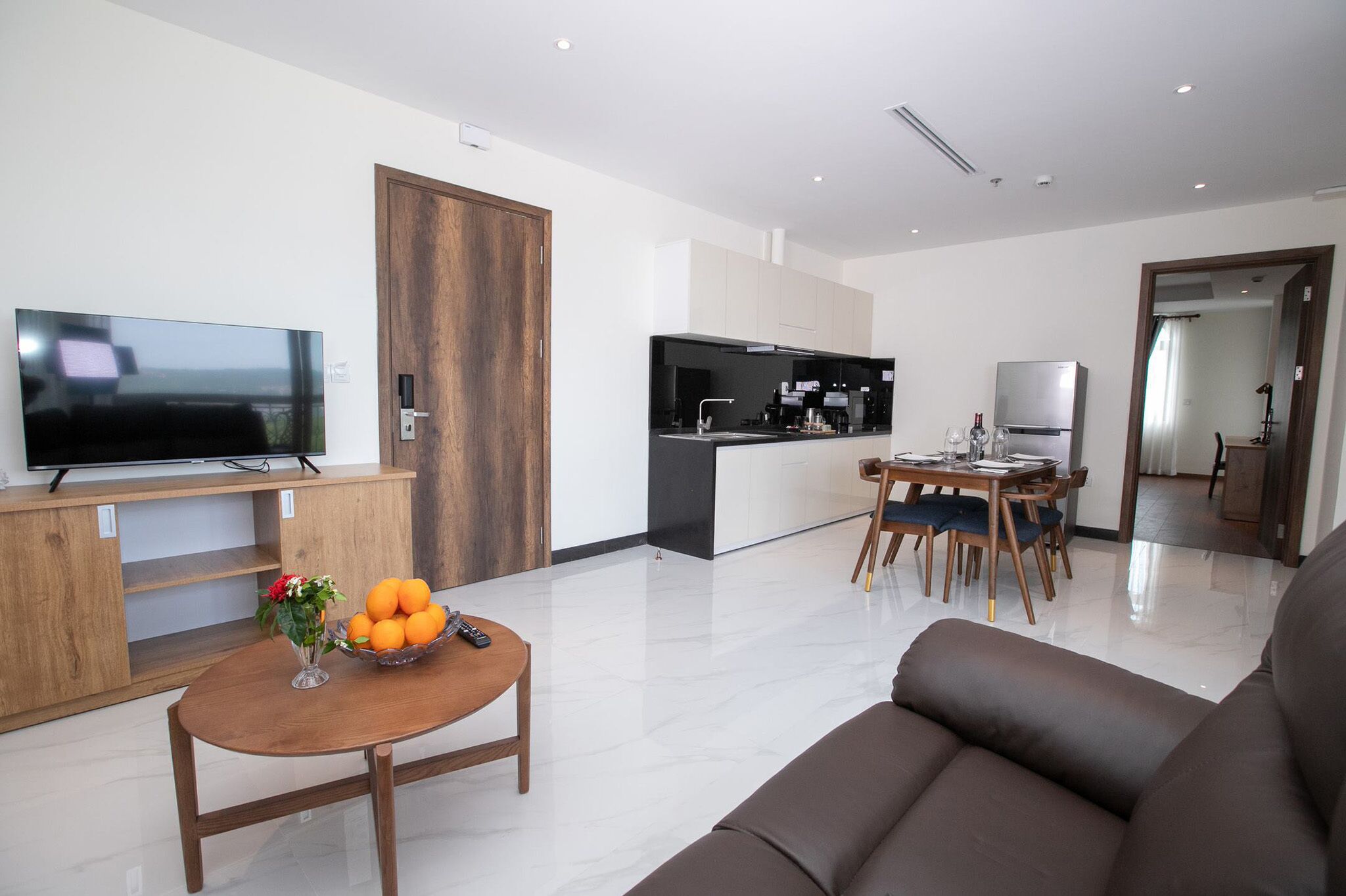 Una sala moderna con un televisor de pantalla plana, una mesa de centro de madera con un florero y naranjas, un sofá marrón y una zona de cocina con nevera y mesa de comedor.