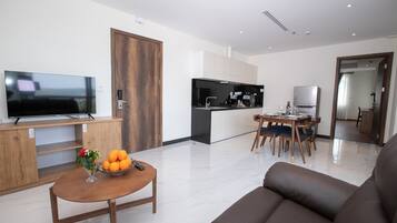Una sala moderna con un televisor de pantalla plana, una mesa de centro de madera con un florero y naranjas, un sofá marrón y una zona de cocina con nevera y mesa de comedor.