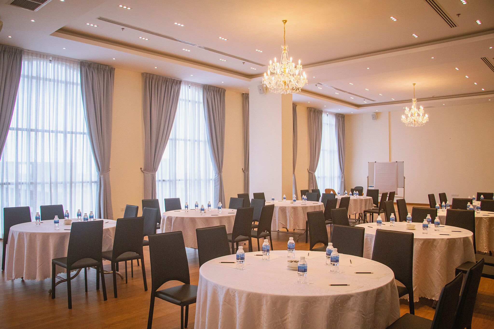 A conference room with multiple round tables, chairs, and water bottles, equipped with a projector and a whiteboard.
