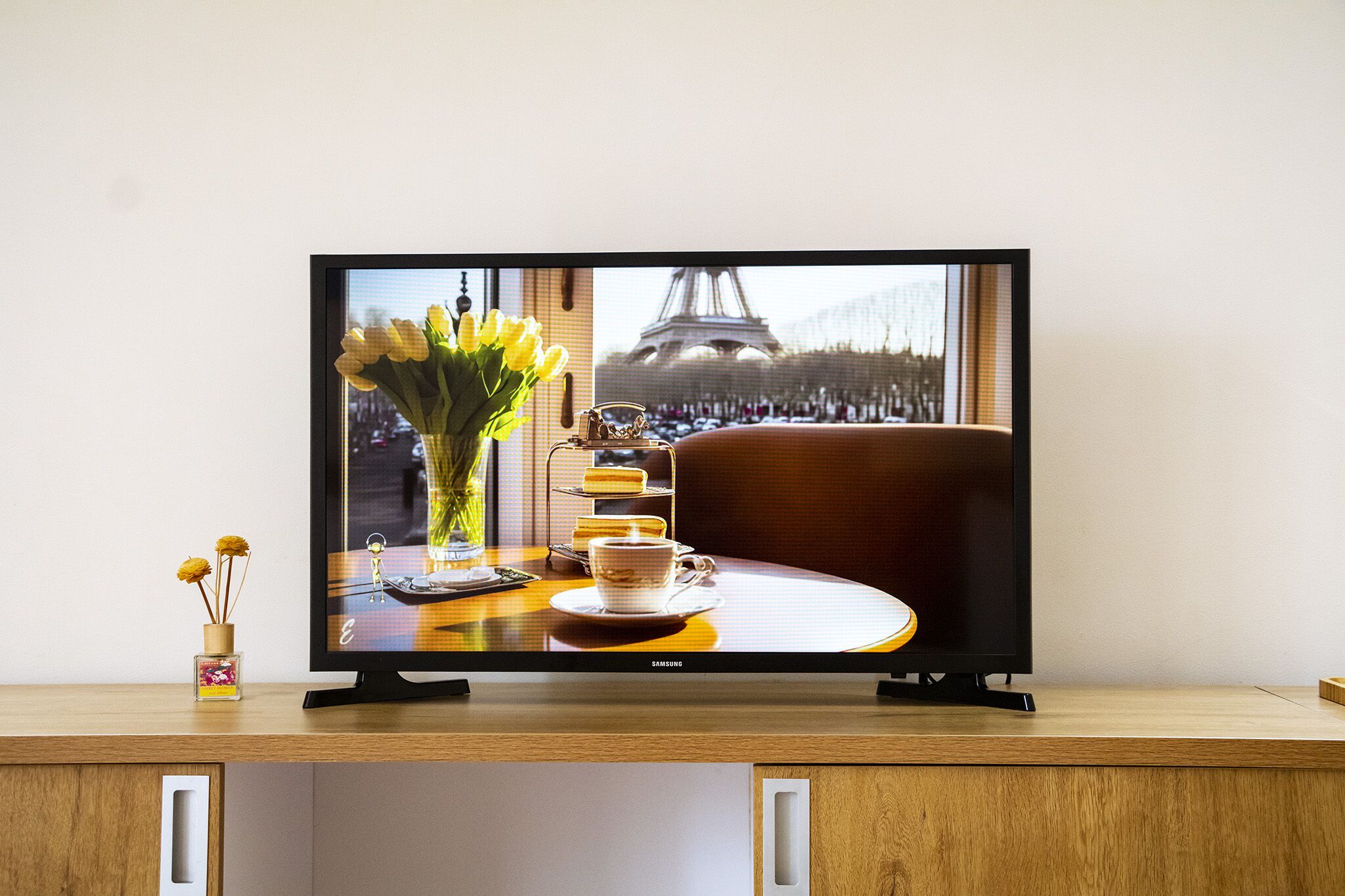 Un téléviseur affiche une scène représentant un vase de fleurs, une tasse et une assiette de desserts sur une table, avec la Tour Eiffel en arrière-plan.
