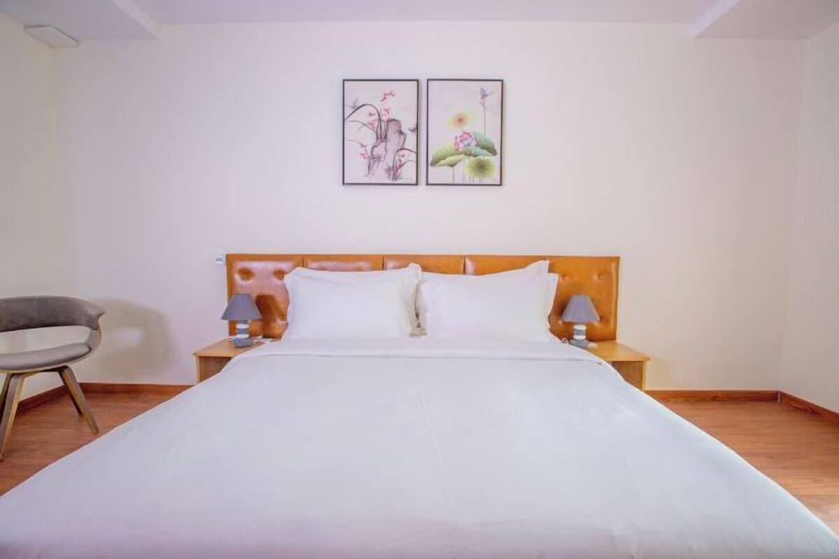 A neatly made bed with white linens, two bedside tables with lamps, a grey chair, and two framed artworks on the wall.