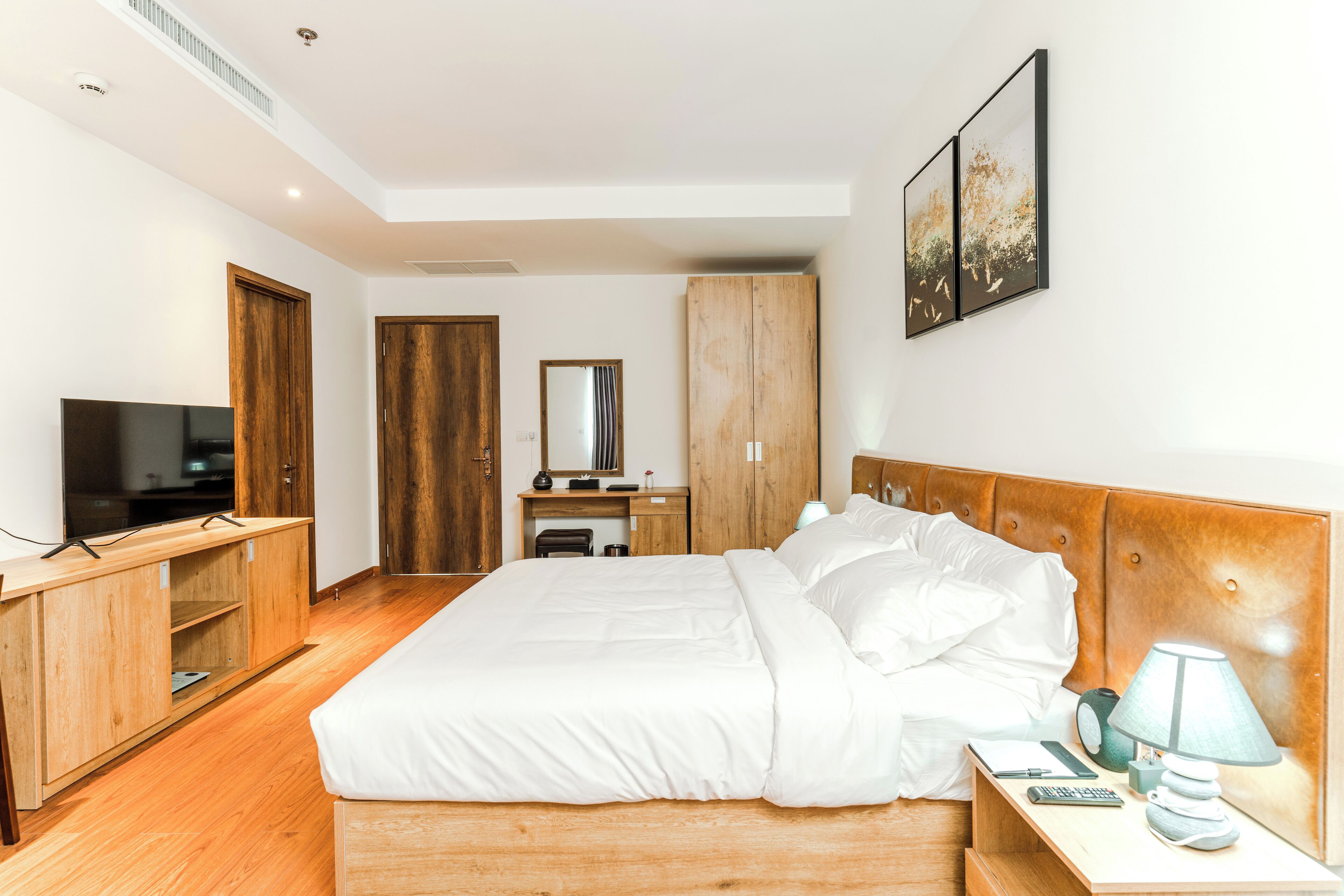 A modern hotel room with a large bed, wooden wardrobe, a desk with a chair, a television, and artwork on the wall.