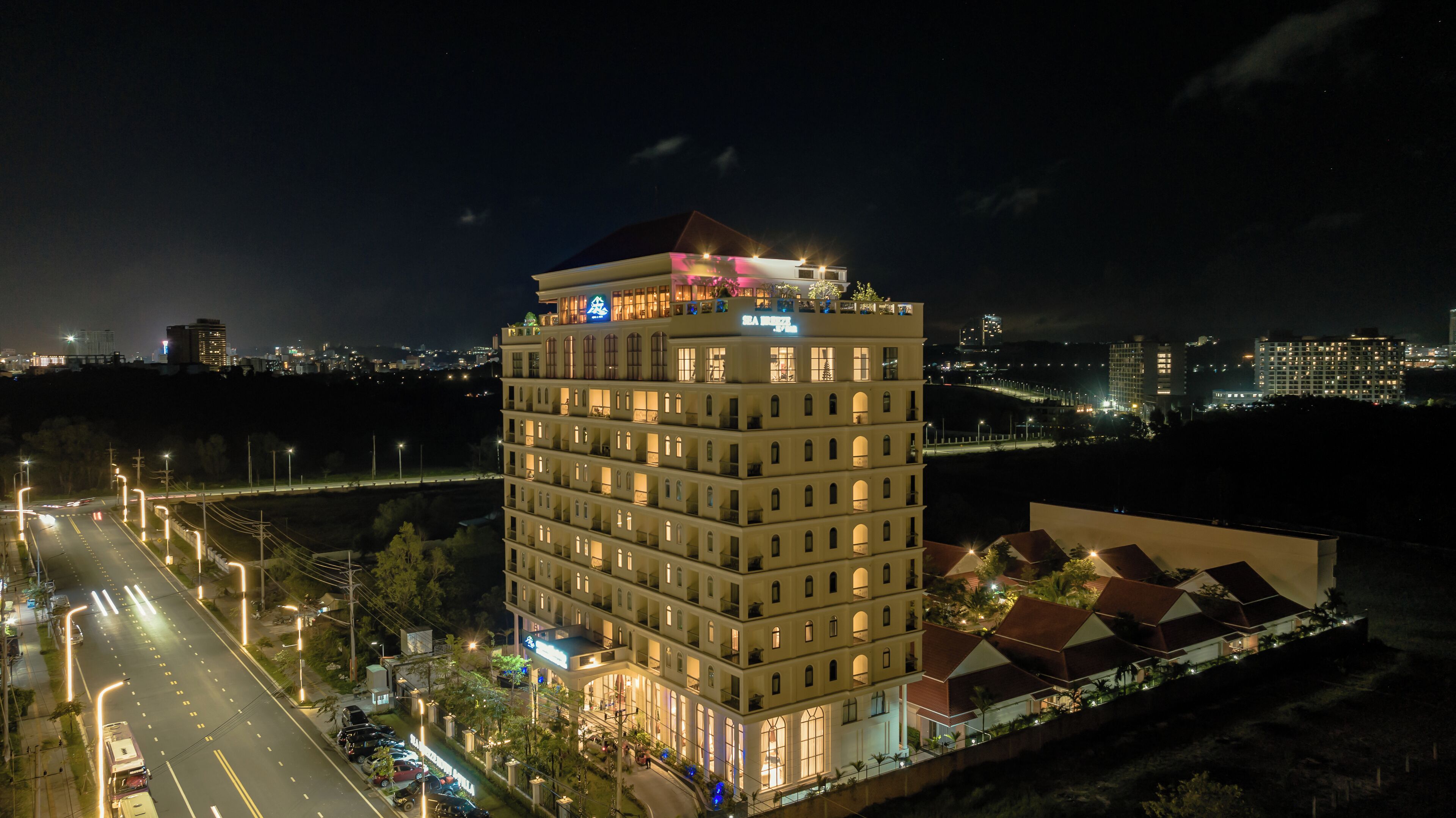 Um hotel com vários andares e um terraço no telhado bem destacado, fachada bem iluminada e vista para a paisagem urbana ao redor.