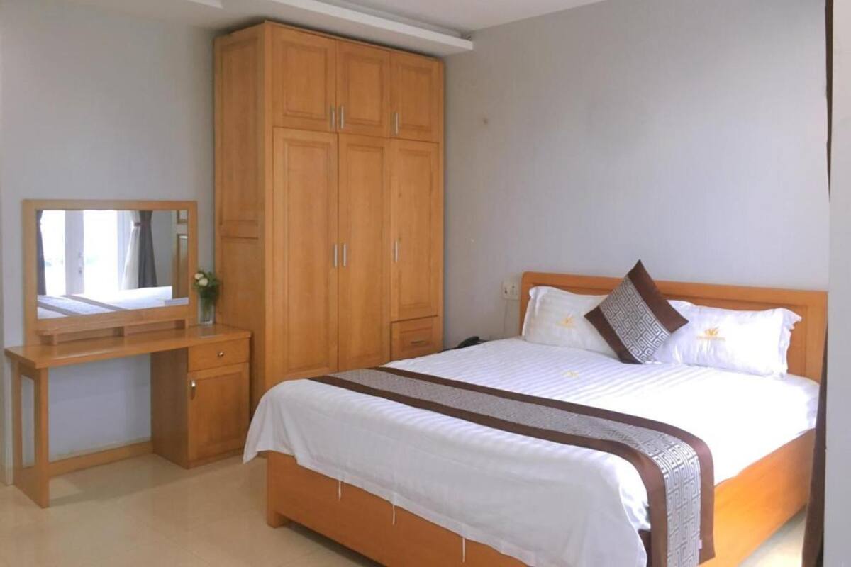 A bedroom with a bed, a wooden wardrobe, a desk with a mirror, and a small vase with flowers.