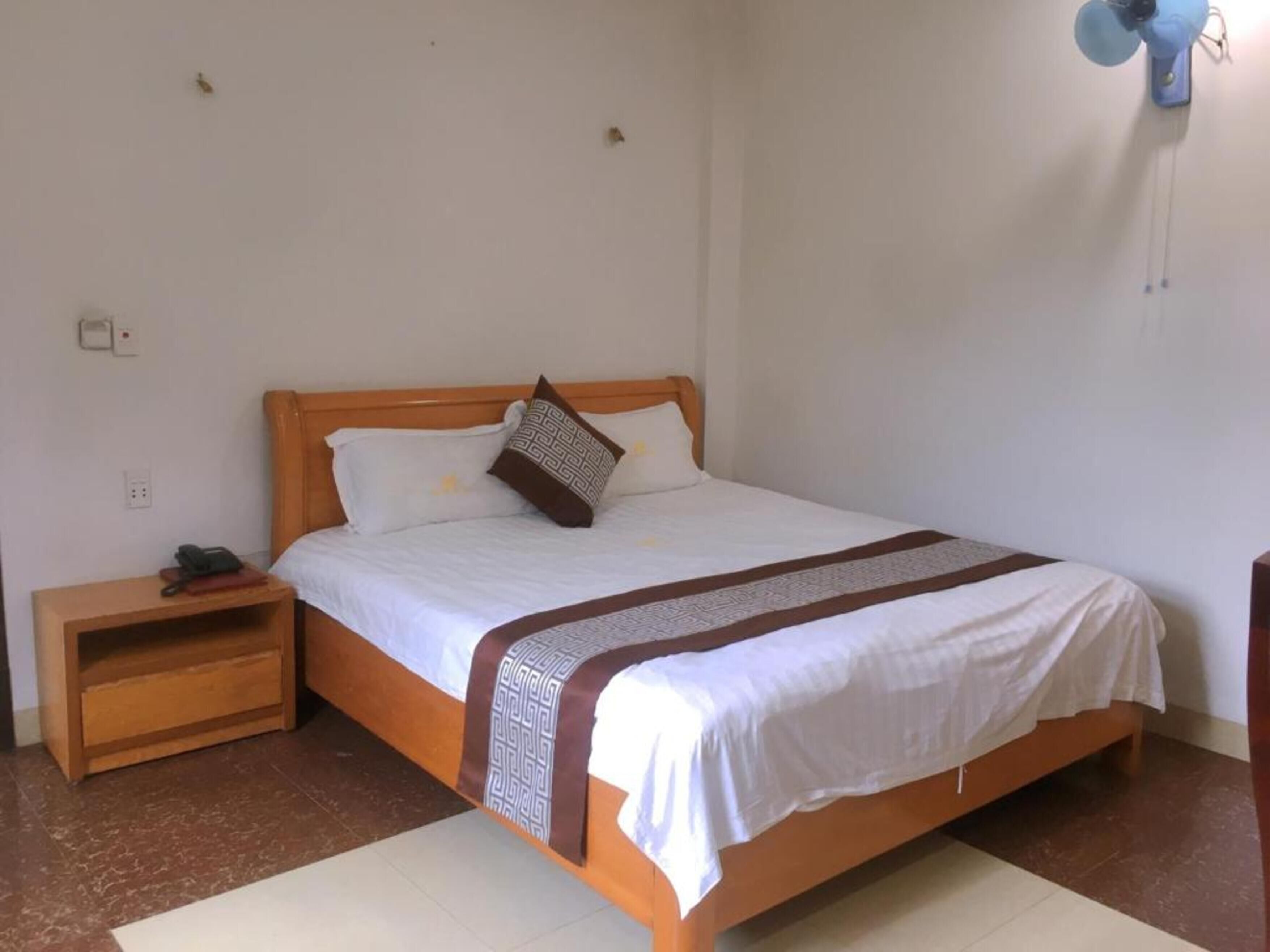A single bed with a wooden frame, white bedding, and a patterned throw. There is a bedside table with a phone and a book, and a wall-mounted fan.