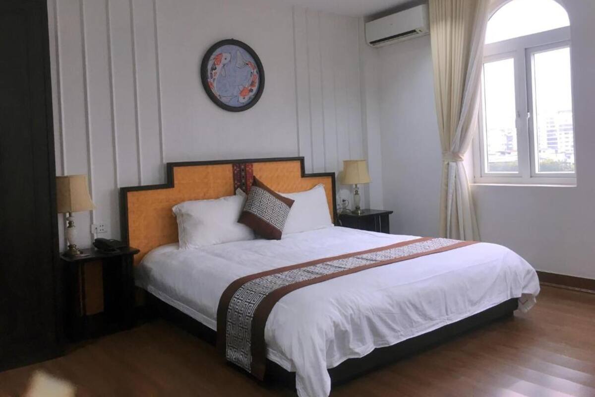 A hotel room with a large bed, two bedside tables, a wall-mounted clock, an air conditioning unit, and a window with curtains.