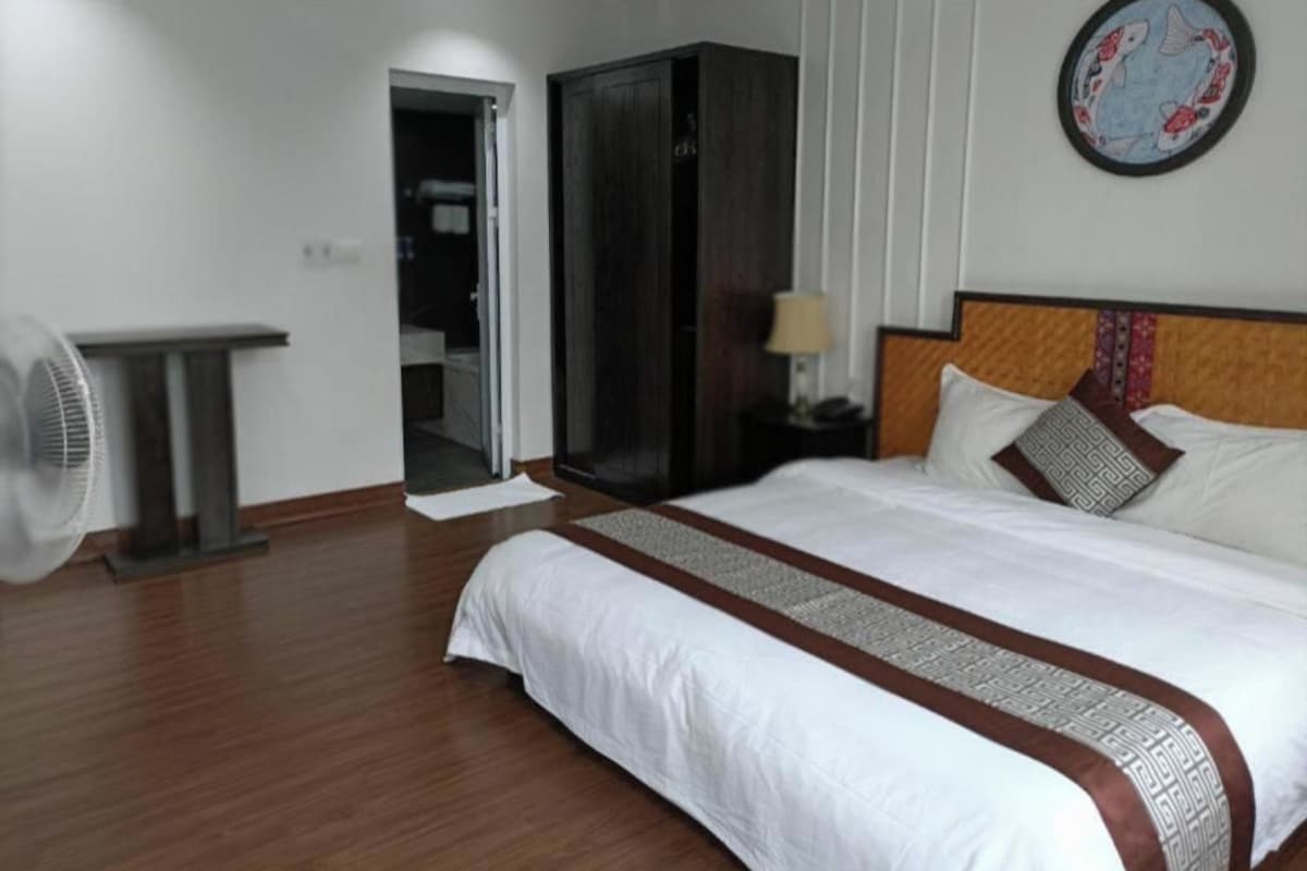 A hotel room with a large bed, a wardrobe, a bedside table with a lamp, and a standing fan.