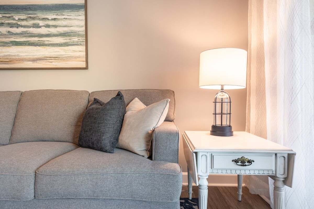 A room with a grey sofa, a white side table with a lamp, and a painting of the ocean on the wall.