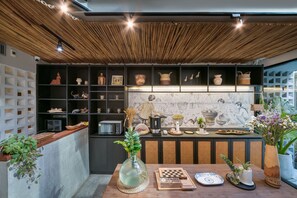 A modern kitchen with a bamboo ceiling, shelves with kitchenware, a dining table with a chess set, and a large window with plants.