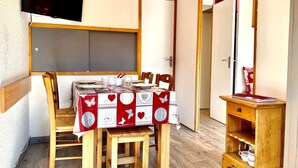 A small dining area with a table set for four, wooden chairs, a television mounted on the wall, and a wooden cabinet with kitchenware.