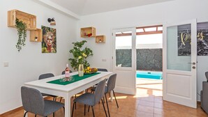 A dining area with a white table, grey chairs, and a green table runner. There are wooden shelves with plants and a painting on the wall. A door leads to a pool area. - Villa Vela Private Pool Playa Blanca Pool Playa Blanca (Playa Blanca)