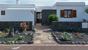 A modern two-story house with a stone facade, a small tower, and a well-maintained garden with various plants and a stone pathway leading to the entrance. - Villa Vela Private Pool Playa Blanca Pool Playa Blanca (Playa Blanca)