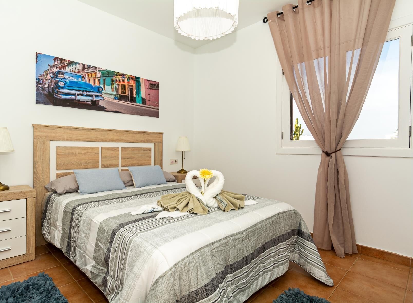 Une chambre bien rangée, avec un lit, une table de chevet, une lampe, une fenêtre avec des rideaux et un tableau accroché au mur.