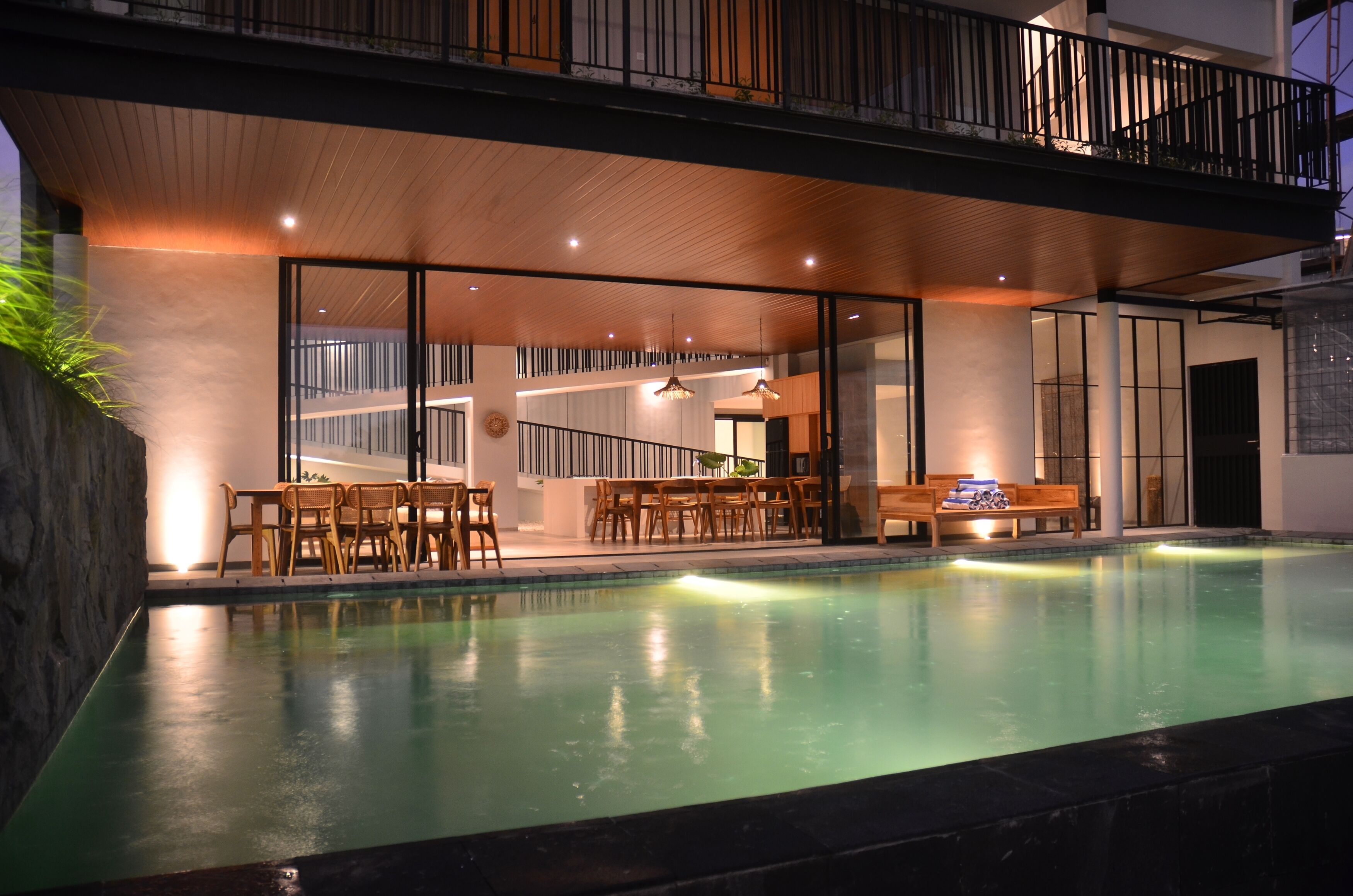 A modern indoor swimming pool with wooden furniture and glass walls, surrounded by a stone wall and a balcony.
