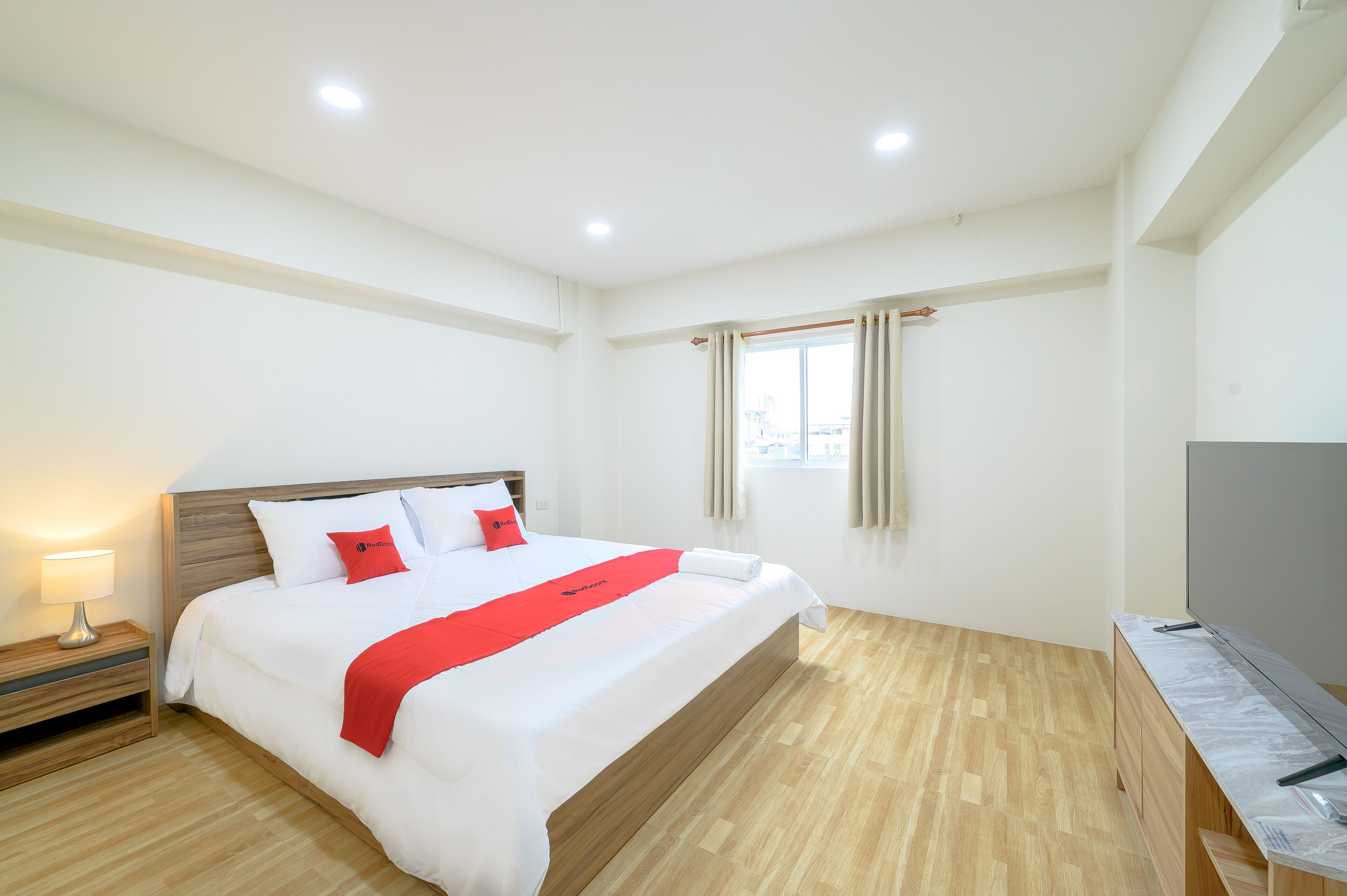 A hotel room with a large bed, a bedside table with a lamp, a TV on a stand, and a window with curtains.